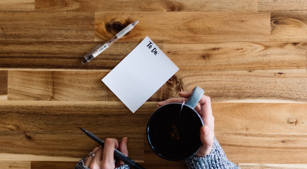 A person writing to-do list on a piece of paper next to a cup of coffee