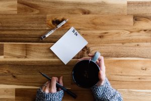 A person writing to-do list on a piece of paper next to a cup of coffee