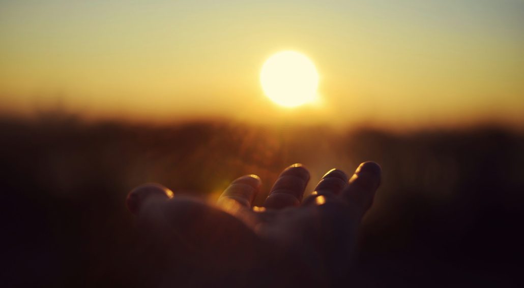 A hand reaching toward the setting sun with warm golden light illuminating the sky