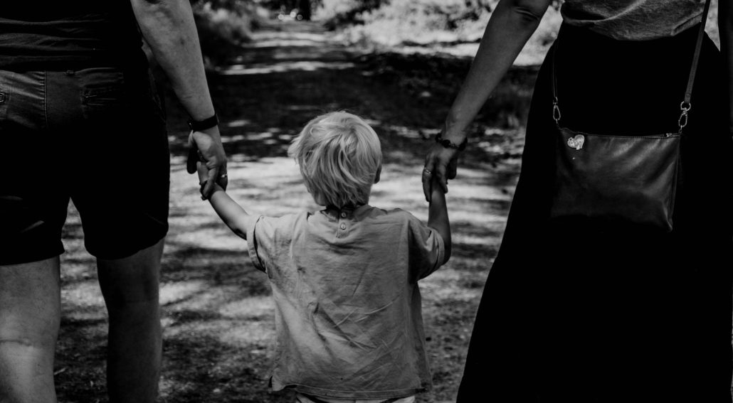 Black and white photo of a small child walking between two adults, holding their hands, on a shaded forest path