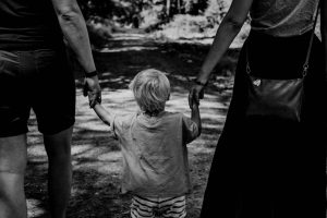 Black and white photo of a small child walking between two adults, holding their hands, on a shaded forest path
