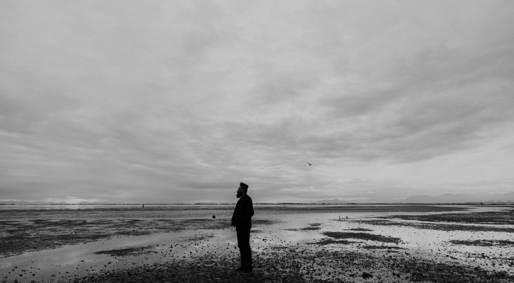 A person standing alone on a vast, reflective shoreline under a cloudy sky, gazing into the distance in a moment of solitude.