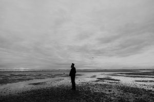 A person standing alone on a vast, reflective shoreline under a cloudy sky, gazing into the distance in a moment of solitude.