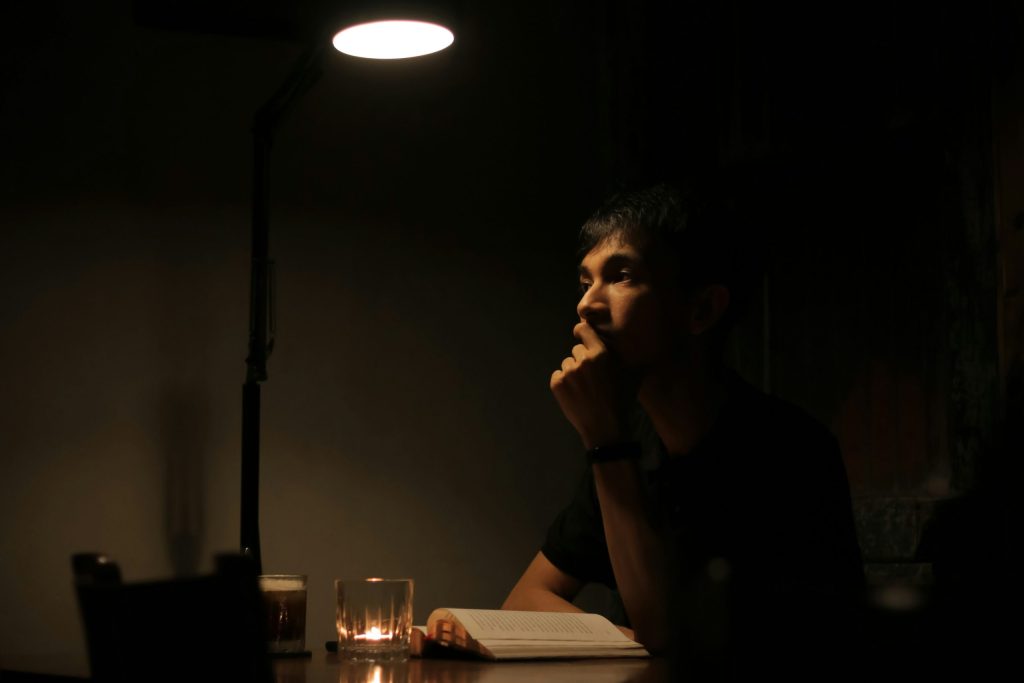 A person sitting alone at a dimly lit table, resting their chin on their hand in deep thought, with an open book and a drink beneath a warm desk lamp.