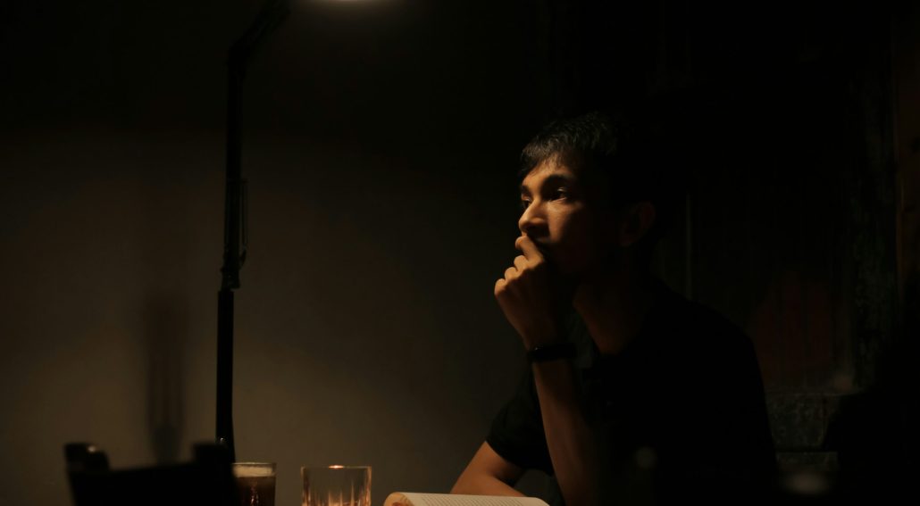 A person sitting alone at a dimly lit table, resting their chin on their hand in deep thought, with an open book and a drink beneath a warm desk lamp.