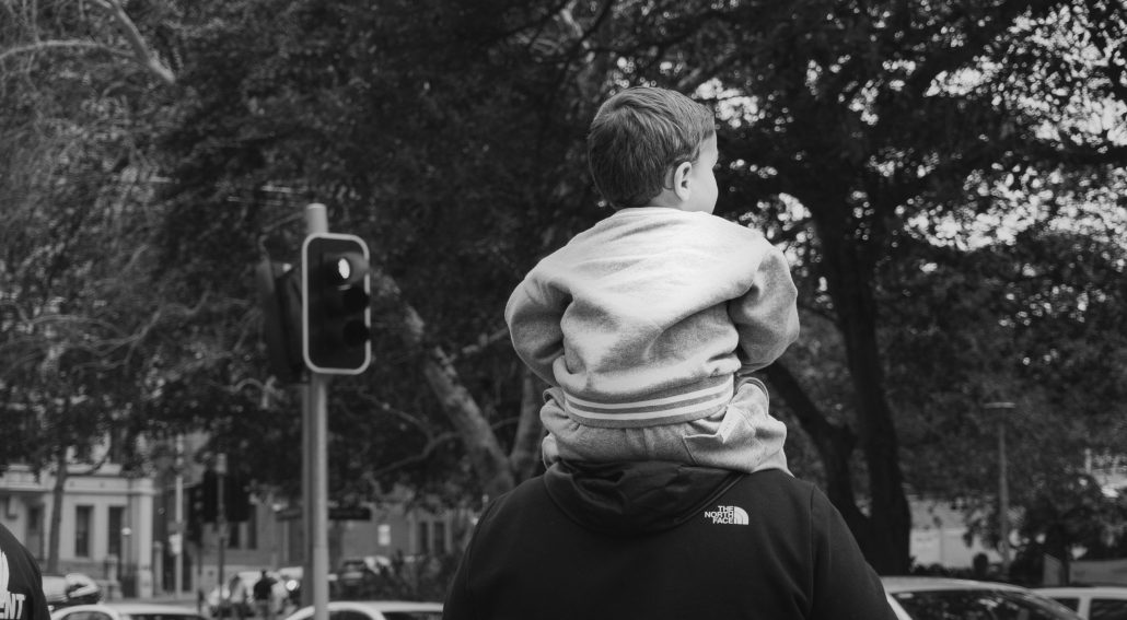Child sitting on an adult’s shoulders at a city crosswalk.