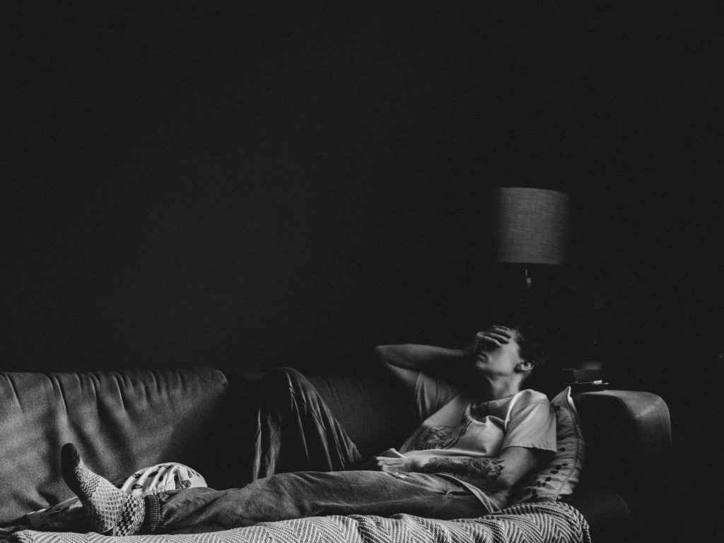 A person lying on a couch with a hand covering their face in a dimly lit room