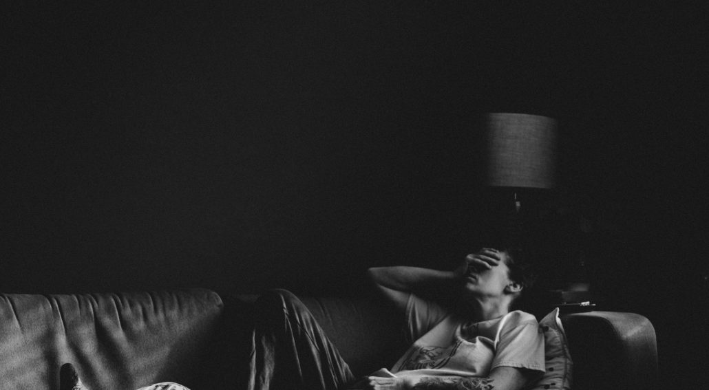 A person lying on a couch with a hand covering their face in a dimly lit room