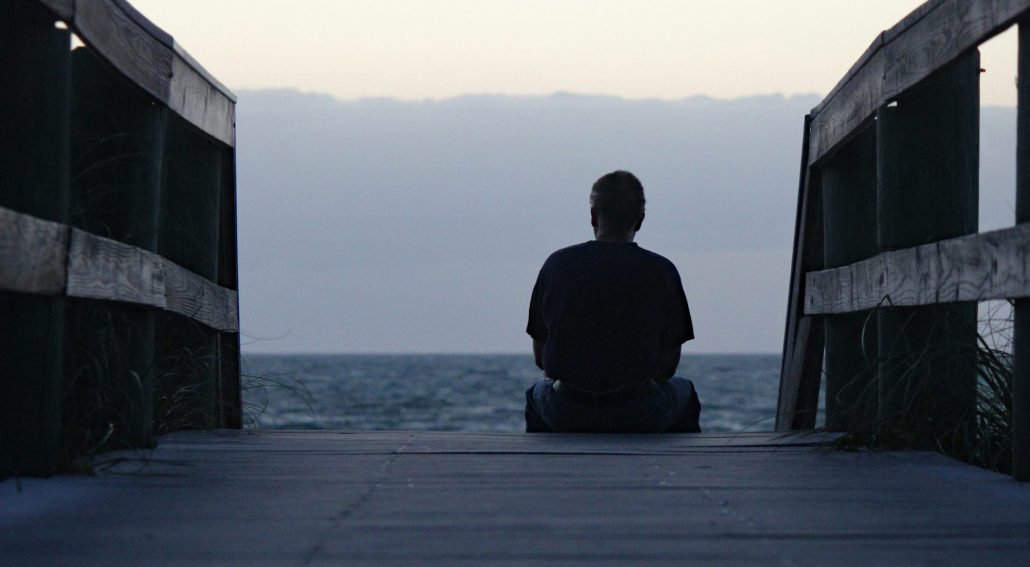 Person sitting at the end of a wooden walkway facing the ocean