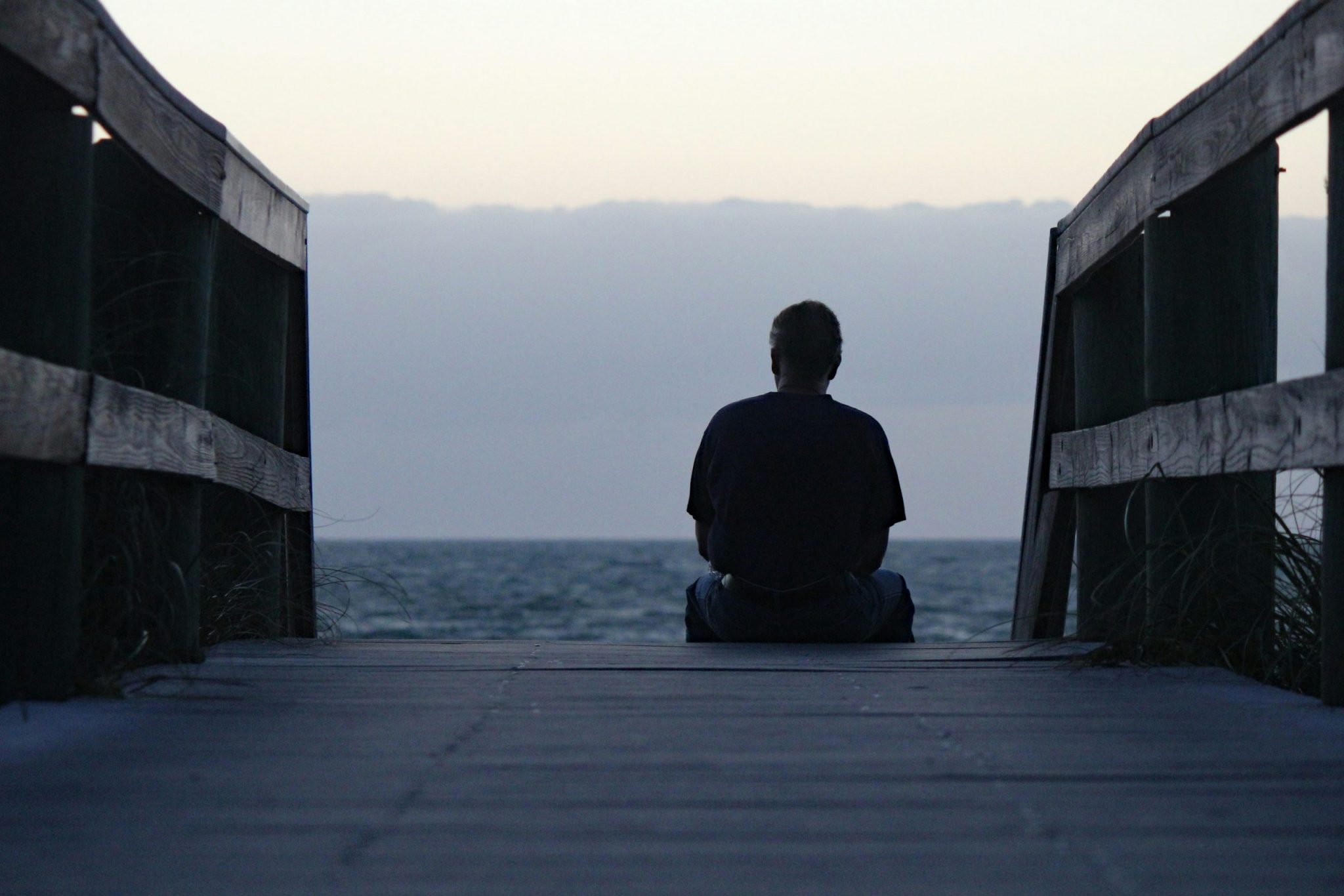 Person sitting at the end of a wooden walkway facing the ocean