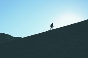 Silhouette of a person walking uphill on a steep ridge against a clear blue sky.