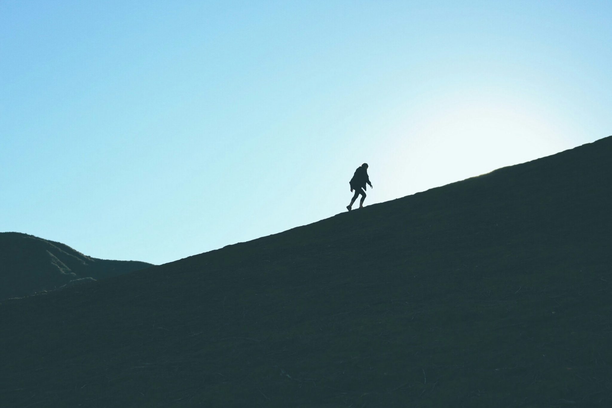 Silhouette of a person walking uphill on a steep ridge against a clear blue sky.