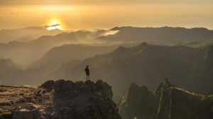 Person standing on a mountain cliff at sunrise, overlooking a misty valley and layered peaks