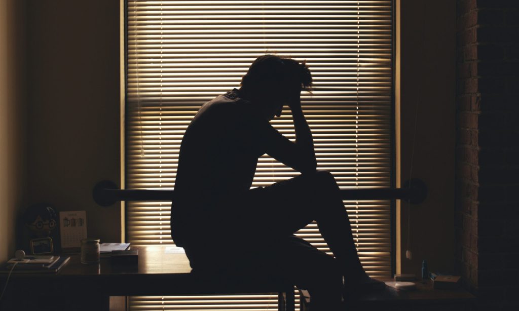 Silhouette of a man inside a room sitting on a table with his hands on his head